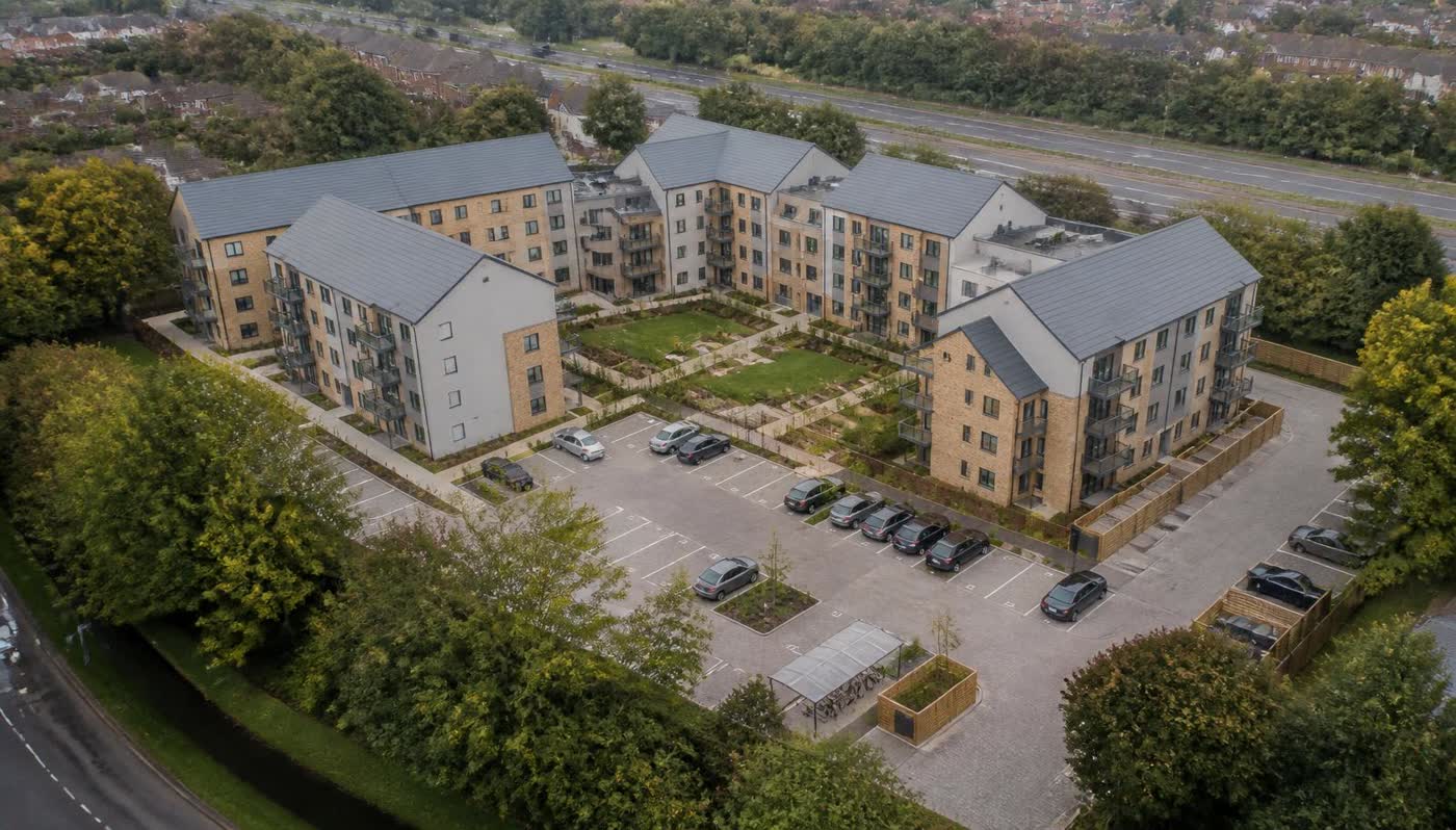 The same site as brick and render apartment blocks with pitched roofs, balconies, a courtyard garden and parking court, same aerial camera position and overcast light